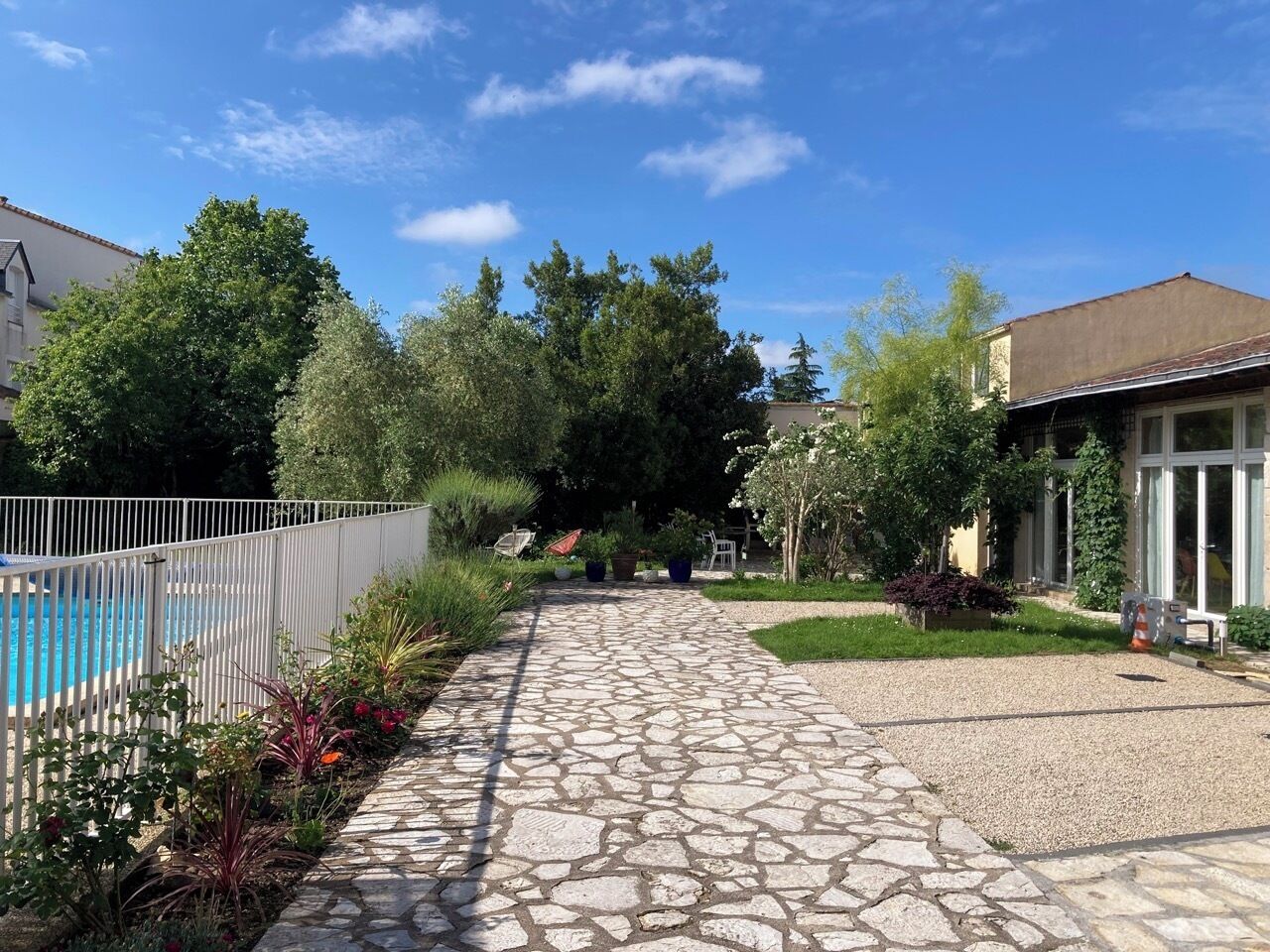 Un sendero de piedra conduce a un área de piscina con piscina cercada, rodeada de vegetación y un edificio con grandes ventanales.
