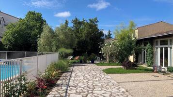 Un sendero de piedra conduce a un área de piscina con piscina cercada, rodeada de vegetación y un edificio con grandes ventanales.