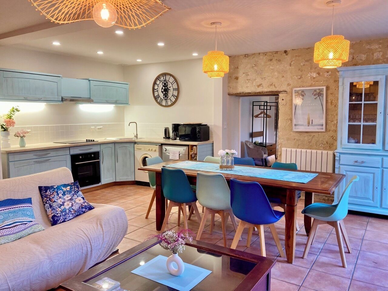 A modern kitchen with light blue cabinets, a dining area with a wooden table and colorful chairs, and a living room with a sofa and a coffee table.