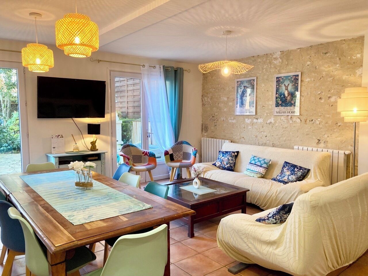 Een gezellige woonkamer met een houten eettafel, twee stoelen, een bank en een televisie. Er staan decoratieve lampen en een wand met twee ingelijste foto’s.