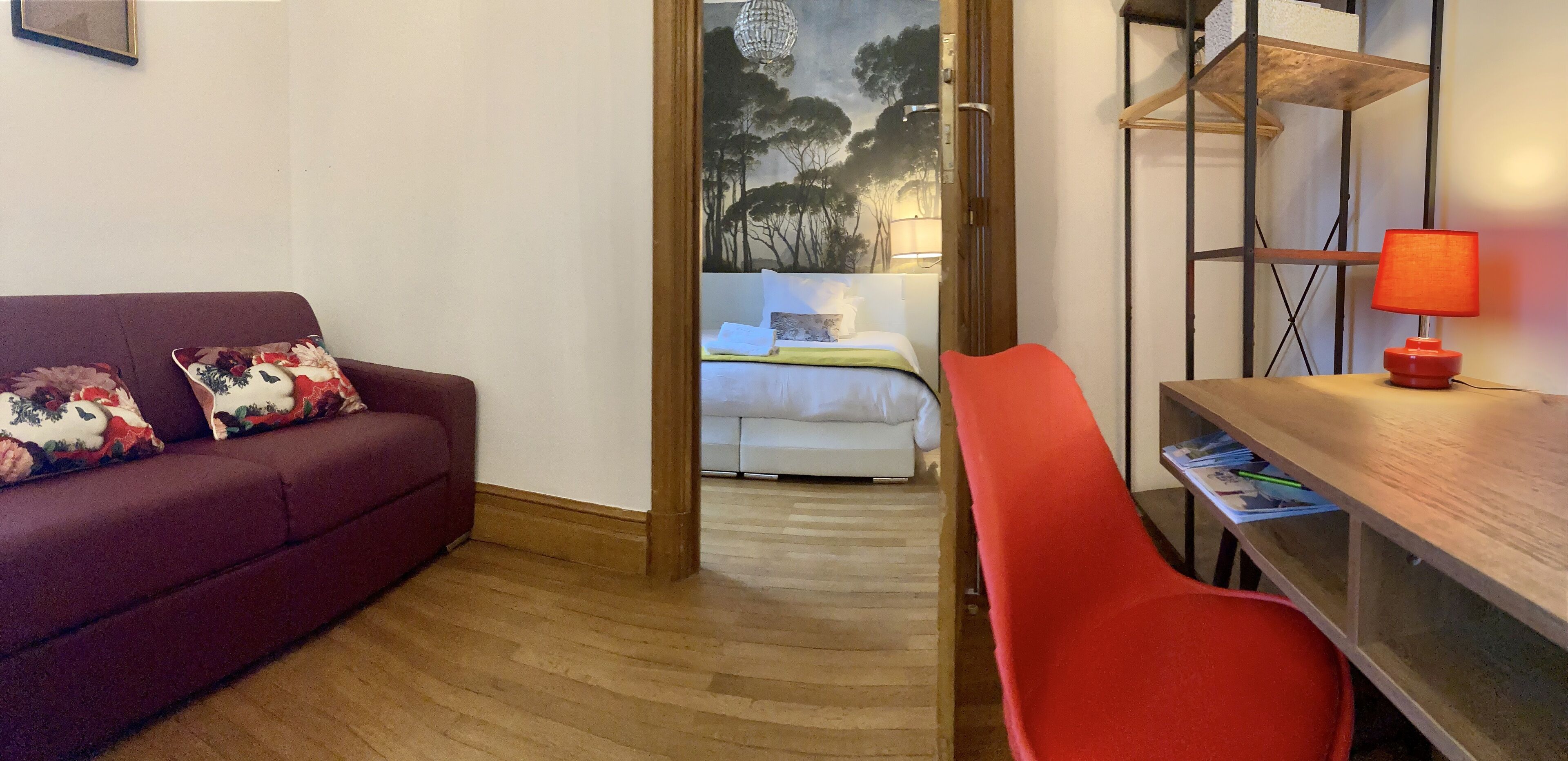 Una habitación con un sofá rojo, un escritorio de madera con una lámpara roja y una cama con cabecero blanco.