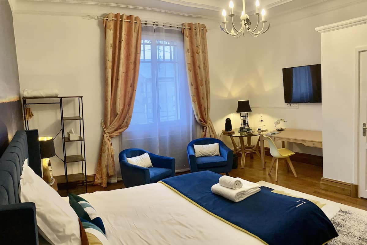 A hotel room with a large bed, two blue armchairs, a desk with a chair, a TV, and a chandelier.