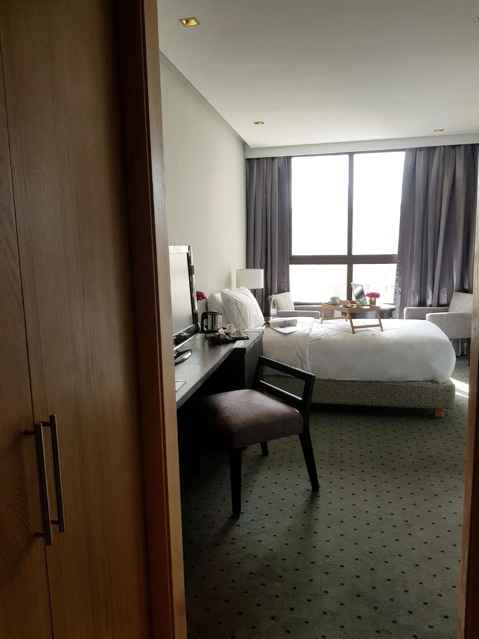 Et hotelværelse med seng, skrivebord, stol, lampe og et vindue med gardiner.