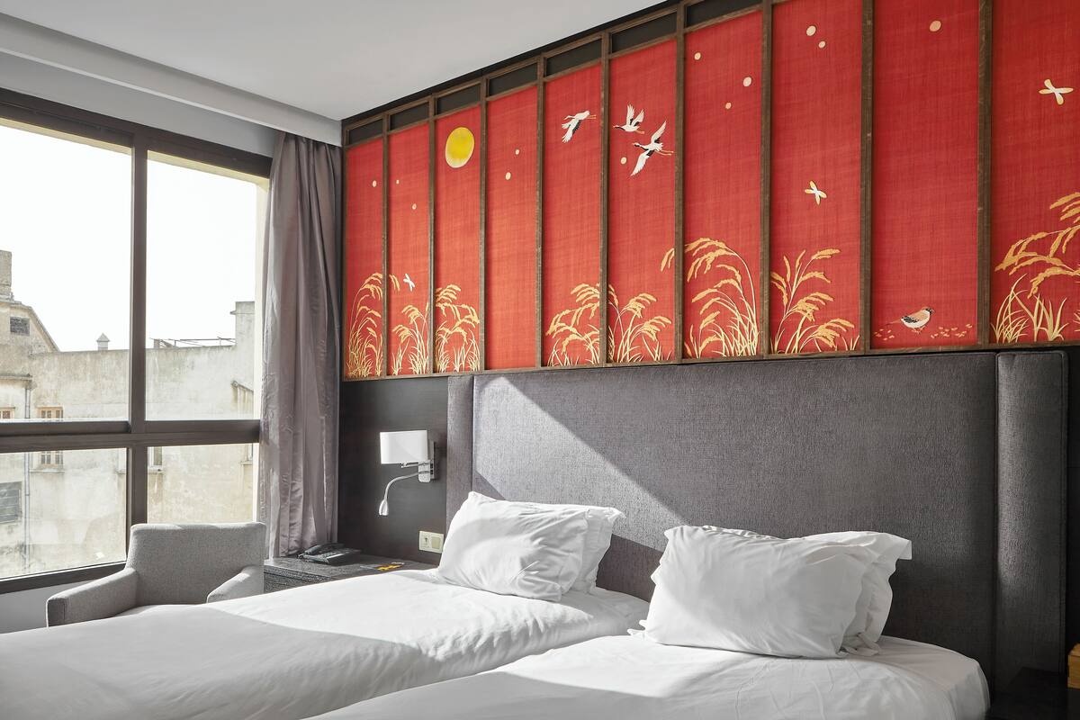 A hotel room with two beds, a large window, and a red decorative panel with birds and plants.