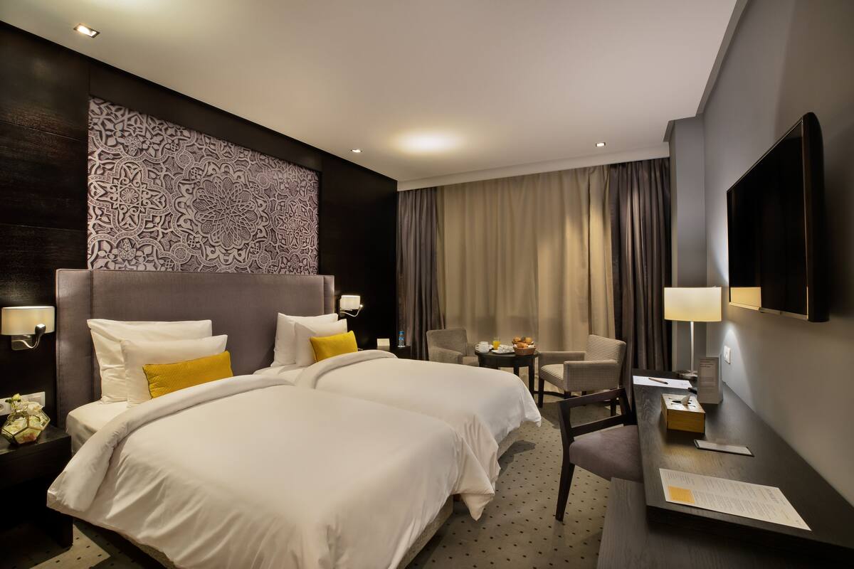 A hotel room with two beds, a desk, a chair, a TV, and a patterned wall.