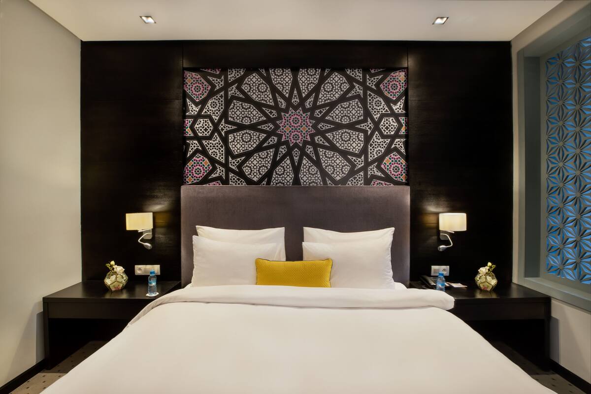 A modern hotel room with a large bed, two bedside tables with lamps, a decorative wall panel, and a patterned window view.