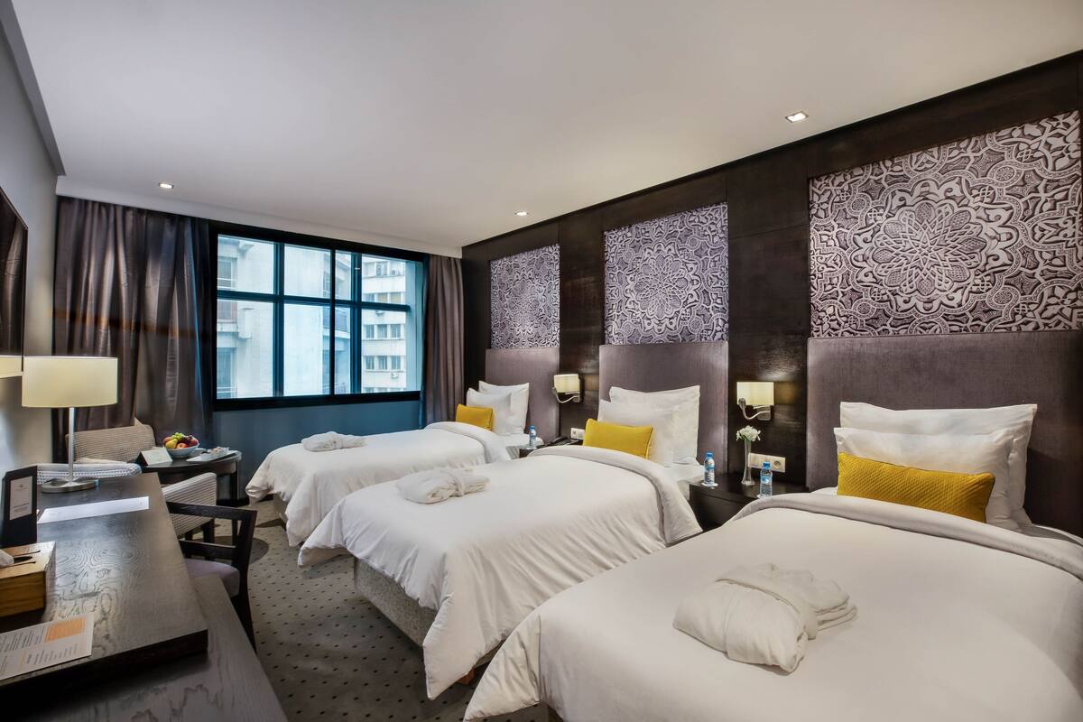 A hotel room with two beds, a desk, a chair, a nightstand, and a large window.