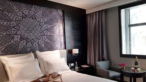 Quarto de hotel com uma cama grande, uma mesa de cabeceira com abajur, uma mesinha com garrafa e copos, uma cadeira e uma janela com cortinas.
