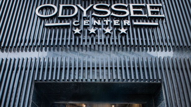A entrada do Odyssey Center com design moderno, apresentando fachada de vidro e um conjunto de escadas que levam à entrada.