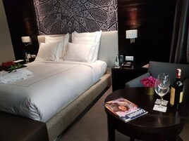 Una habitación de hotel con una cama bien hecha, una mesita de noche con una revista, una botella de vino y una copa.