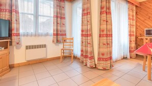 Una habitación acogedora con techo de madera, televisor sobre un mueble, una silla, una mesa y cortinas estampadas.
