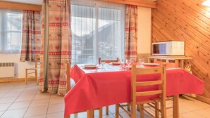 Un comedor con mantel rojo, sillas de madera, un microondas sobre una mesita y cortinas estampadas.