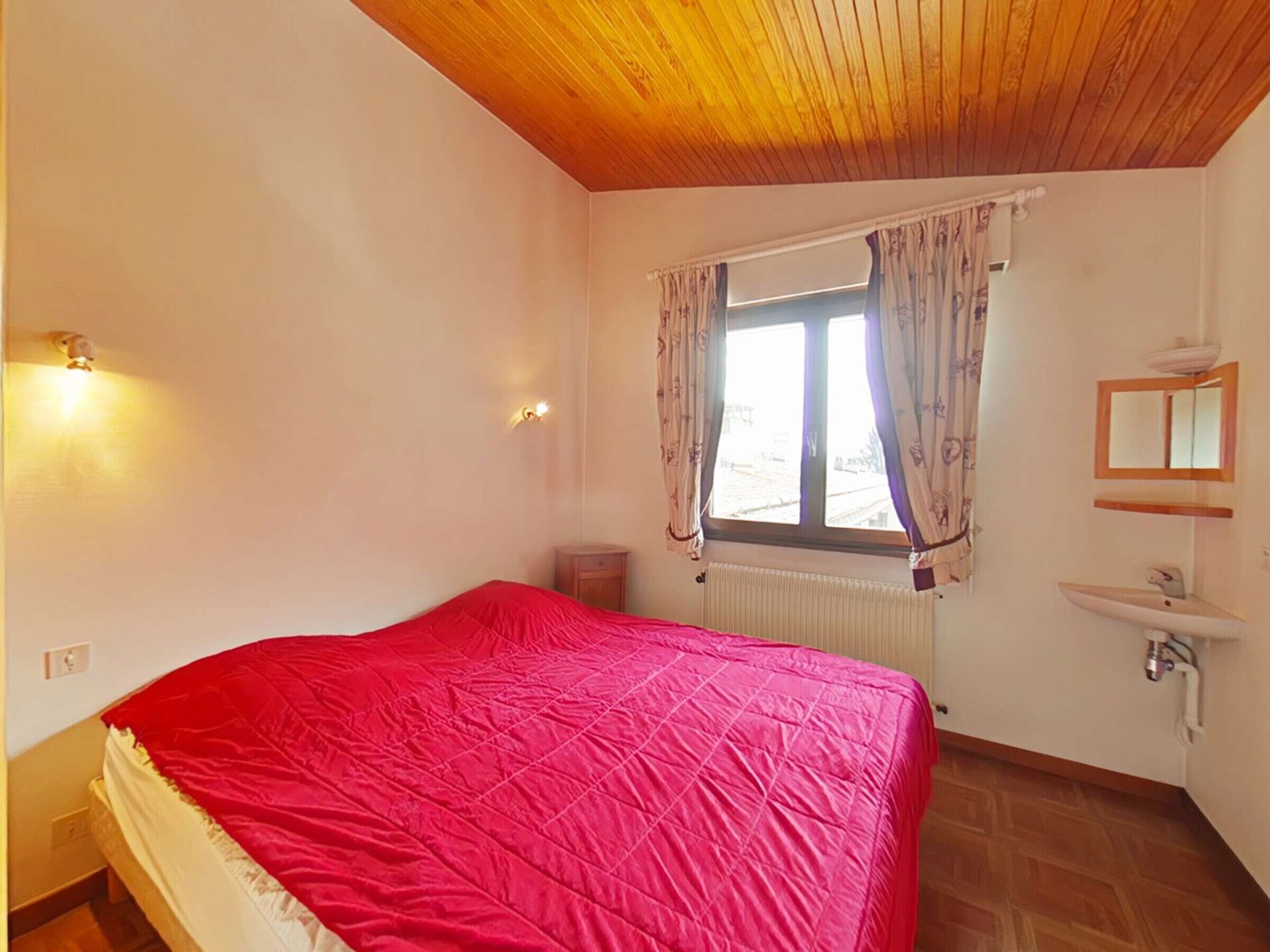 A bedroom with a bed, a window with curtains, a bedside table, a radiator, and a wall-mounted sink.