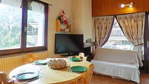 A cozy room with a round wooden table set for two, a flat-screen TV mounted on the wall, a window with curtains, and a sofa with a white cover. - Sunny 2BR Mezzanine Apt with Mountain Views, Fireplace, Near Shops & Lifts, Sleeps 6 (Le Grand-Bornand)