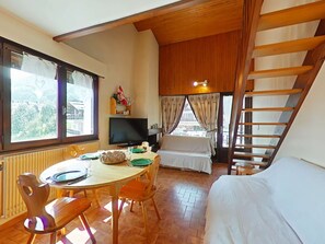 Ein gemütliches Zimmer mit Holzbalkendecke, einem Esstisch mit Stühlen, einem Bett, einem Fernseher und einem Fenster mit Vorhängen.