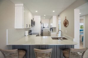 A modern kitchen with white cabinetry, a central island, stainless steel appliances, and a view into the living area. - Come Stay at Beautiful Gulf Front Castle Beach Condo 302 on Fort Myers Beach! (Fort Myers Beach)
