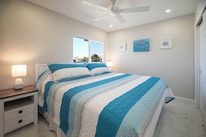 A bedroom with a bed, a nightstand, a lamp, a ceiling fan, and a window with a view. - Come Stay at Beautiful Gulf Front Castle Beach Condo 302 on Fort Myers Beach! (Fort Myers Beach)