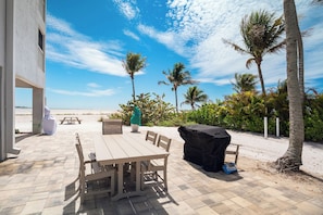 An outdoor patio with a table and chairs, a barbecue grill, and a view of the beach and palm trees. - Come Stay at Beautiful Gulf Front Castle Beach Condo 302 on Fort Myers Beach! (Fort Myers Beach)
