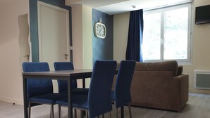 A dining area with a table and chairs, a brown sofa, a clock on the wall, and a window with curtains. - Le Bercail (Saint-Jean-de-Monts)