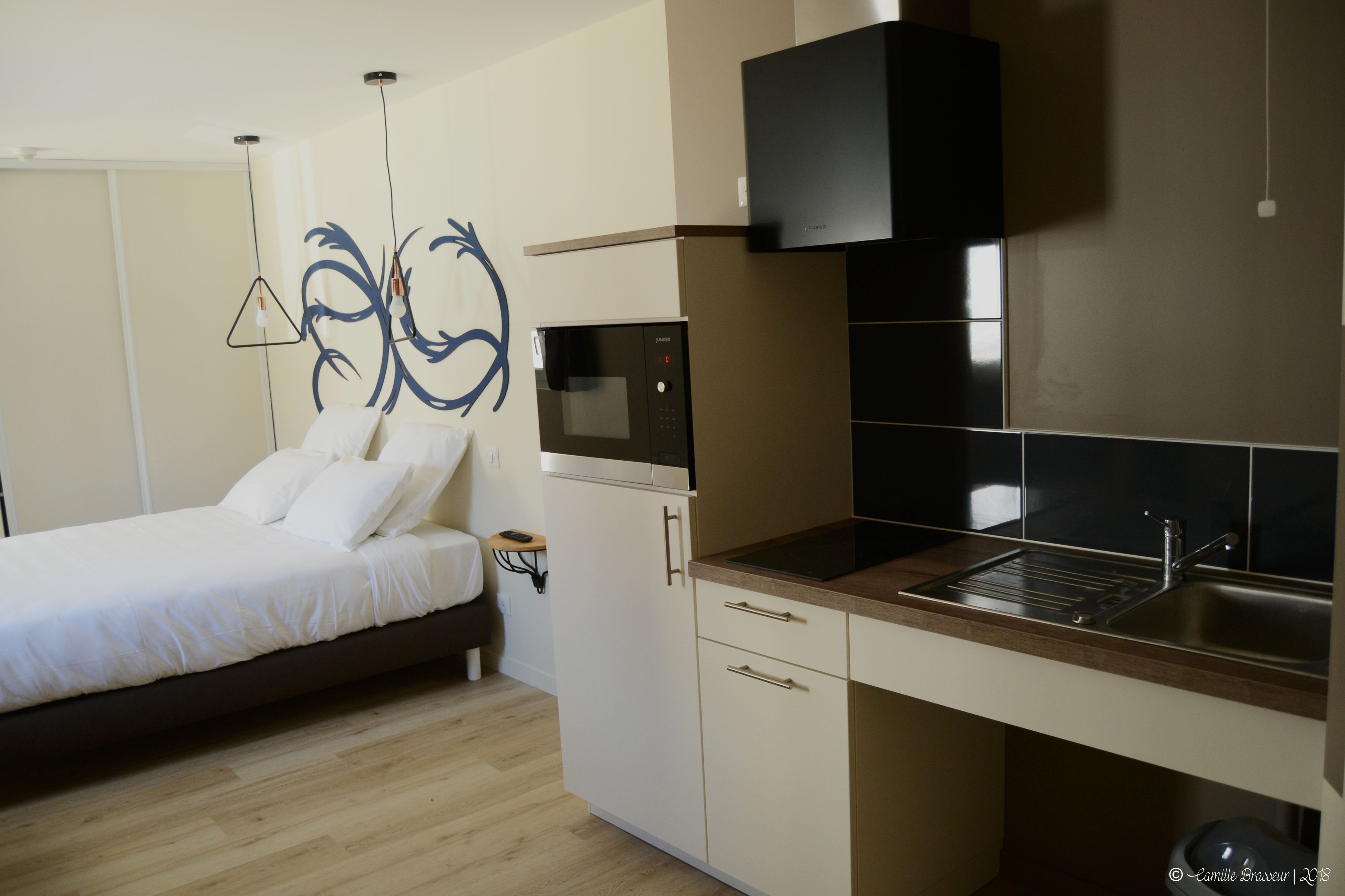 A modern bedroom with a bed, bedside table, and a wall-mounted artwork. Adjacent is a kitchenette with a microwave, stove, and sink.