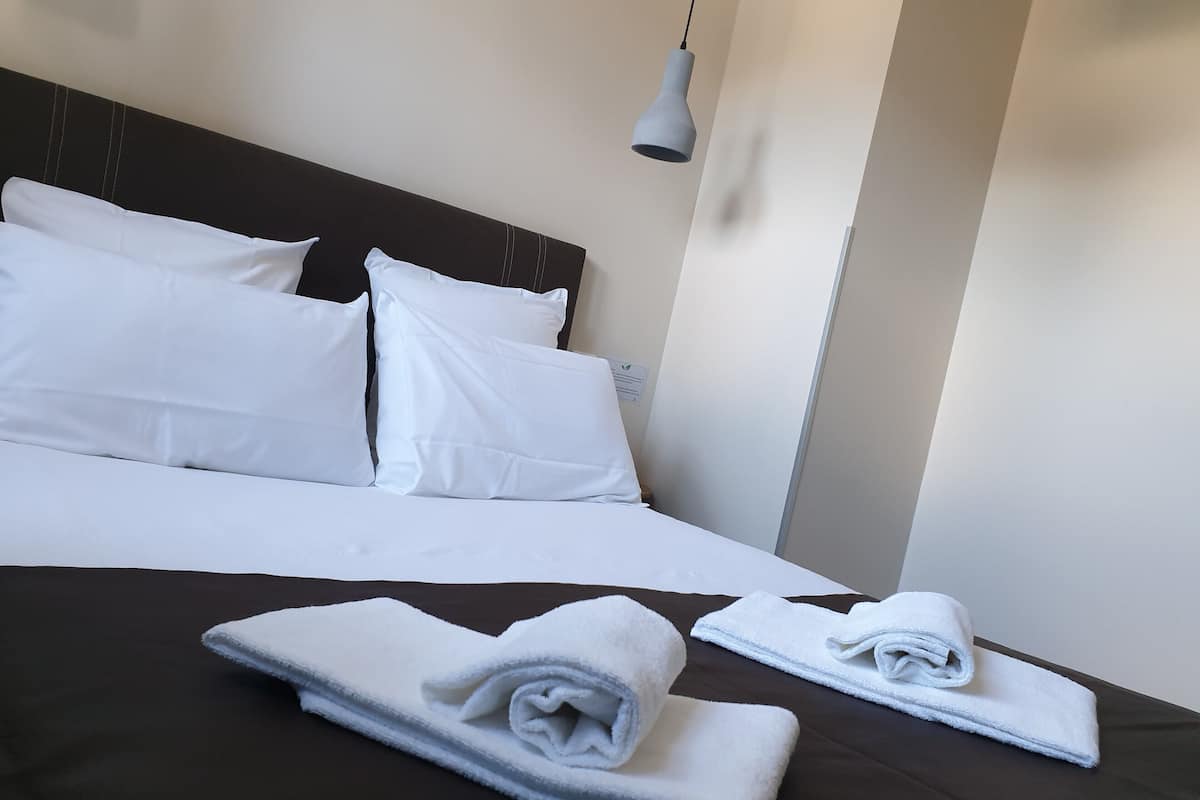A neatly made bed with white linens and two rolled towels on a dark brown bedspread.