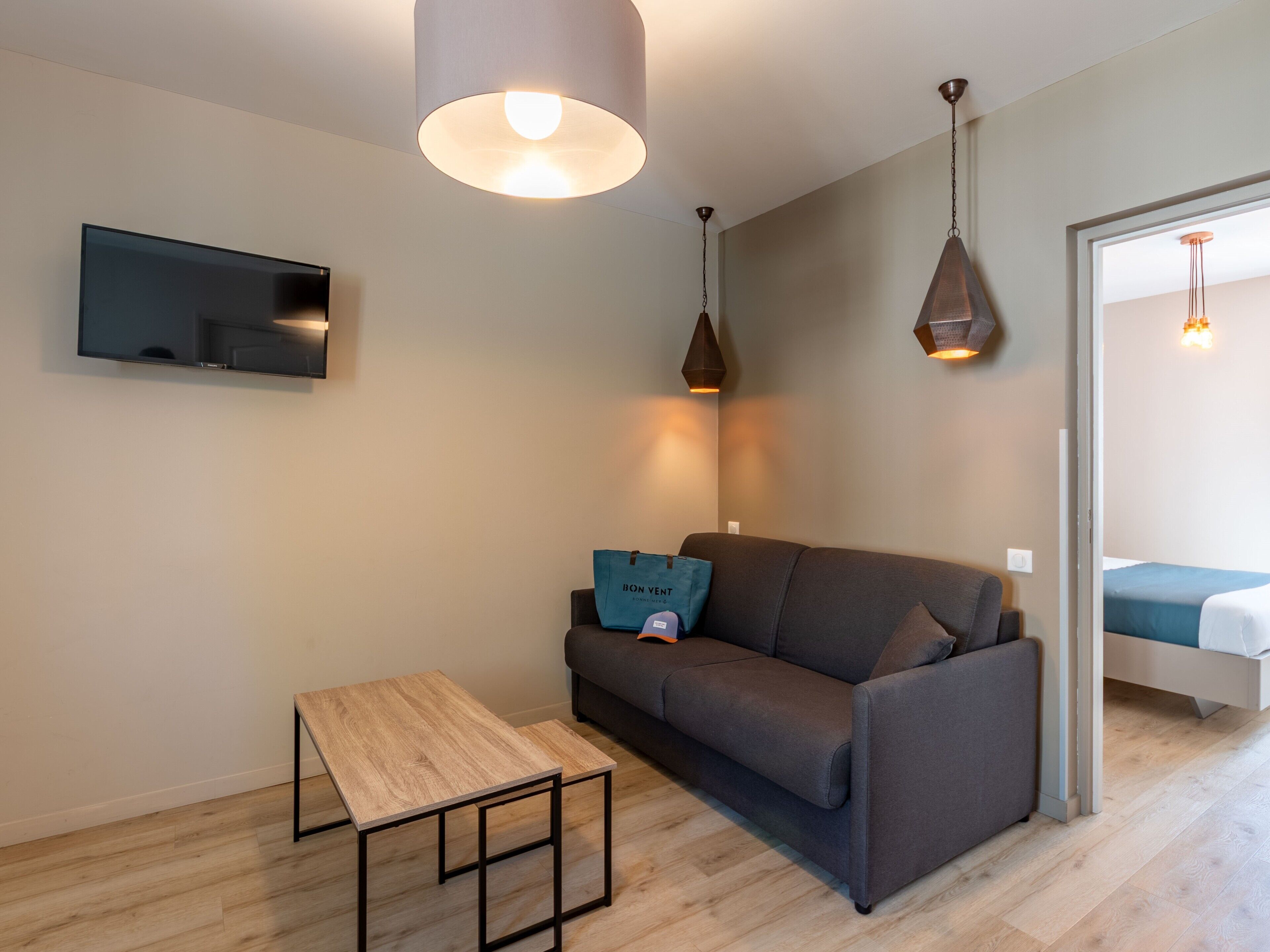 Una sala moderna con un sofá, una mesa de centro, un televisor de pantalla plana y lámparas colgantes, y una cama en la habitación contigua.