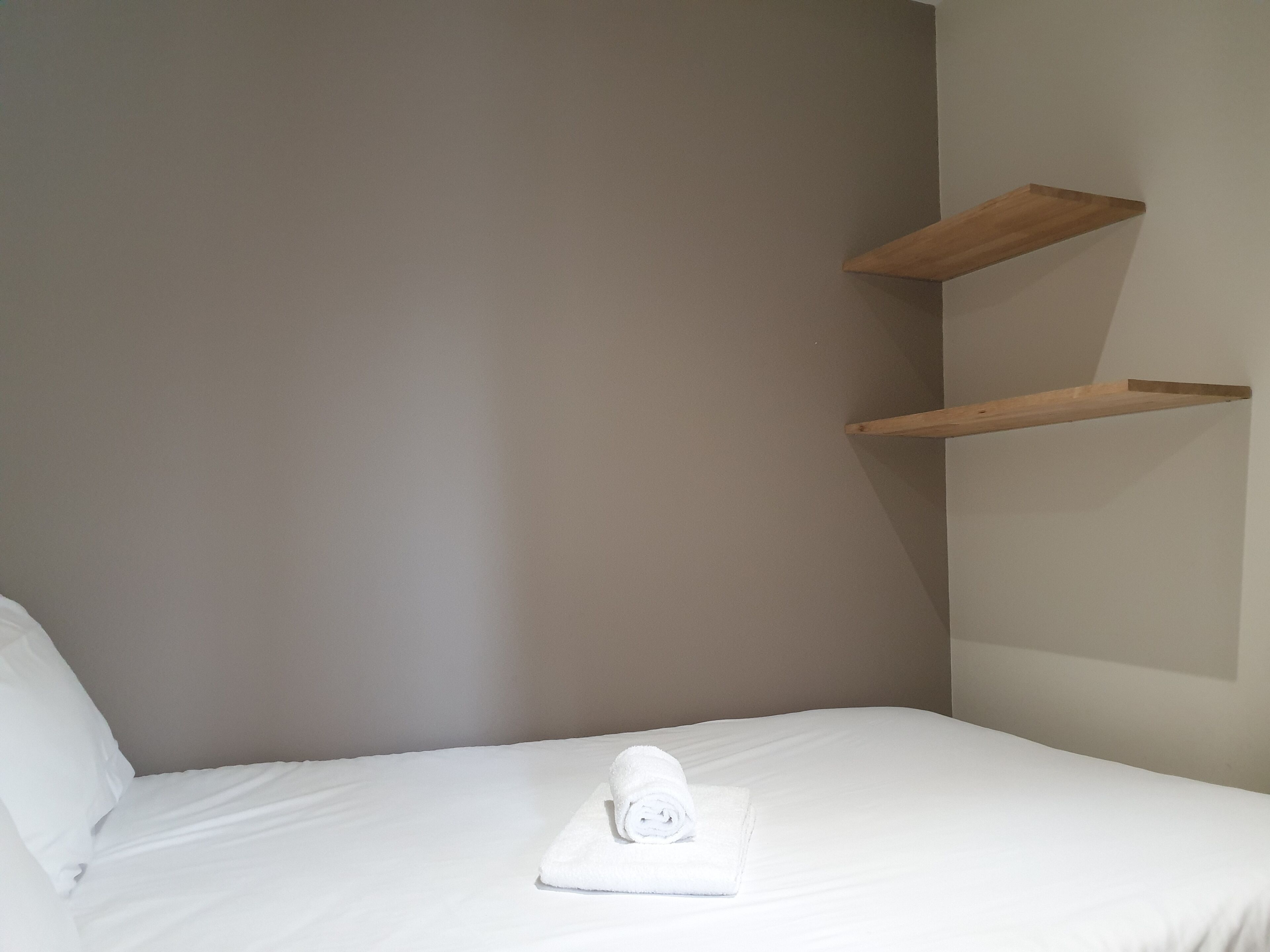 A single bed with a white pillow and a rolled white towel on it. There are two wooden shelves attached to the wall.