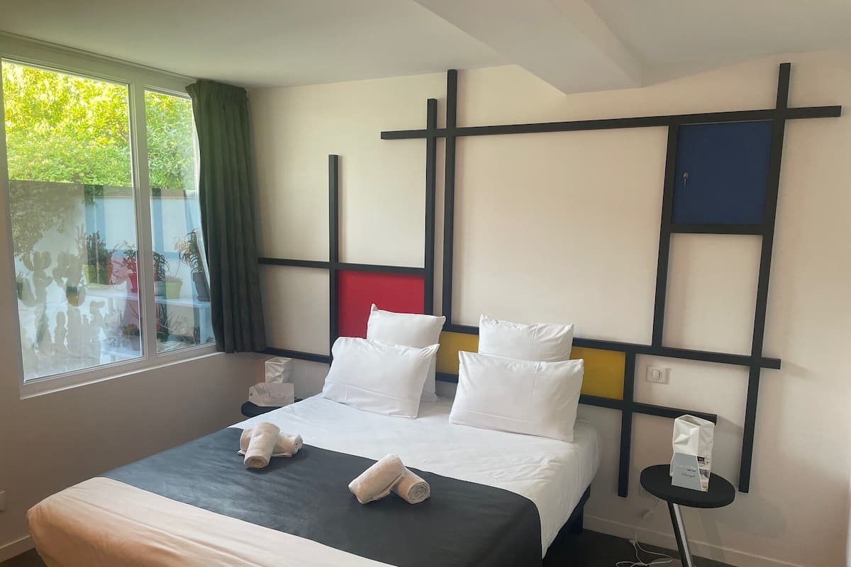 A hotel room with a bed, a bedside table, a window with curtains, and a view of greenery outside.