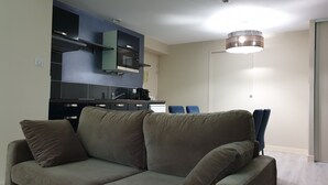 A modern living room with a grey sofa, a kitchen area with dark cabinets, and a ceiling light fixture. - Le Bercail (Saint-Jean-de-Monts)