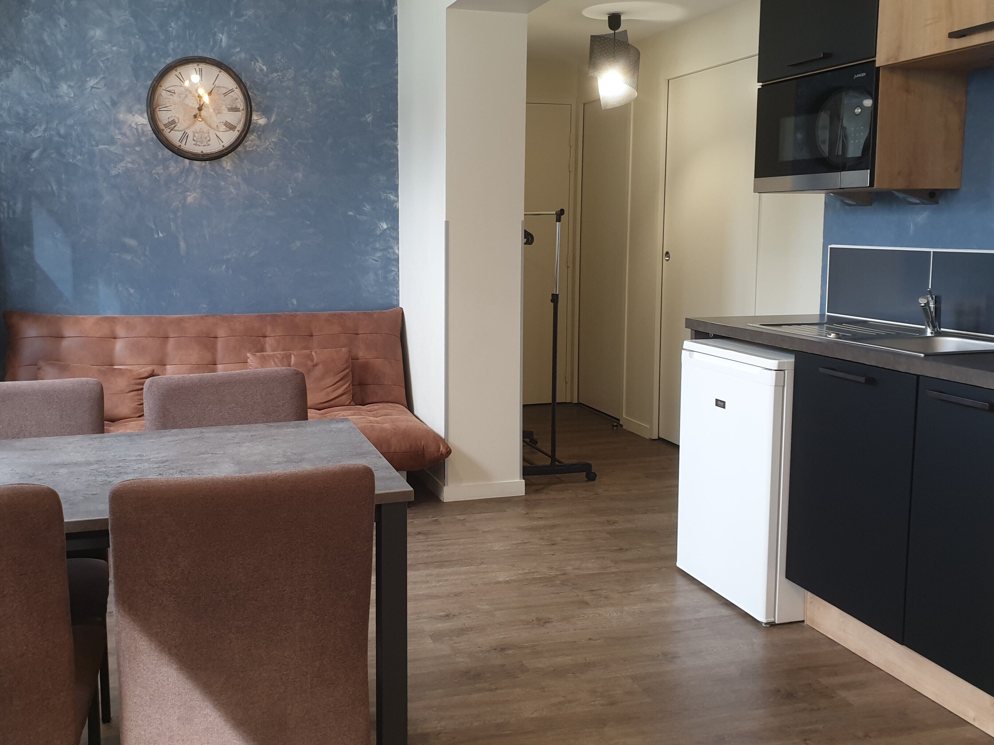 Een moderne keuken met een vaatwasser, een eethoek met een tafel en stoelen, en een blauwe accentmuur met een klok.