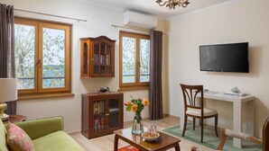 A living room with a sofa, a coffee table, a TV, and a desk with a chair. There are windows with curtains, a cabinet, and a vase with flowers. - Valdibora Seaview Residence (Rovinj)