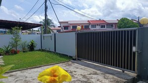 A gated residential area with a concrete pathway, a yellow flower in the foreground, and houses with red roofs in the background. - H Homestay - Semi D 1000Mbps (1Gbps) Wifi, Astro & Private Parking (Sibu)