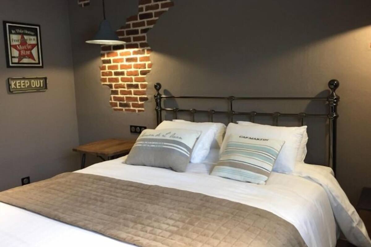 A neatly made bed with a quilted cover, two pillows with text, a metal headboard, a wall-mounted light, and a 'KEEP OUT' sign.