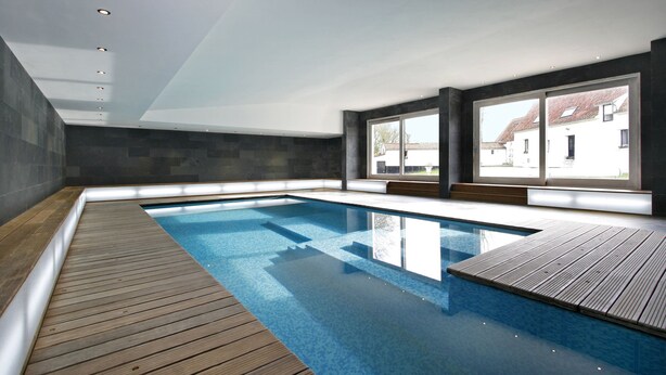 A modern indoor swimming pool with a wooden deck, large windows, and a view of a white building. - Le Clos Du Miroir (Courset)