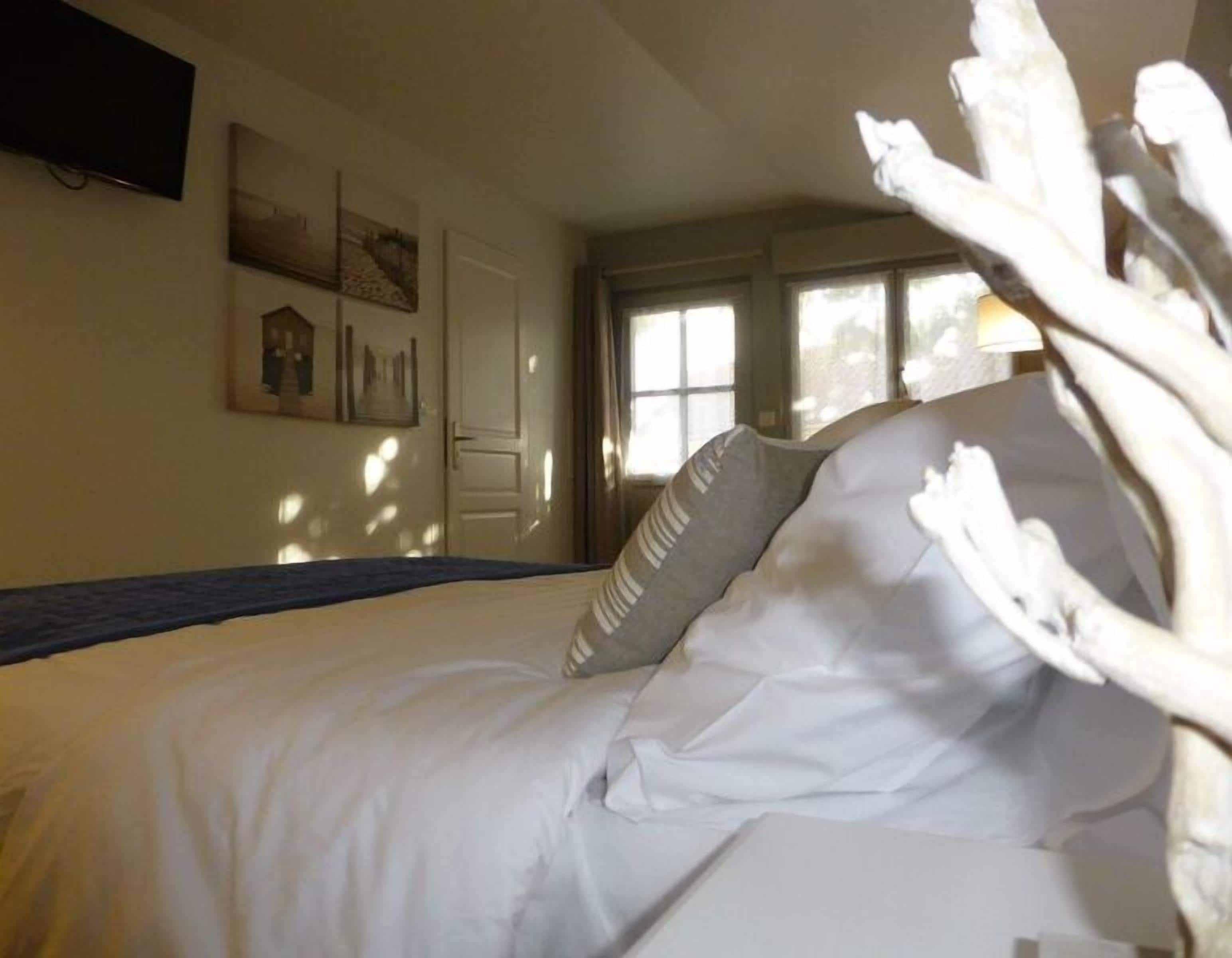 Una cama bien tendida con sábanas blancas y almohadas a rayas, un televisor de pantalla plana montado en la pared y una ventana con cortinas.