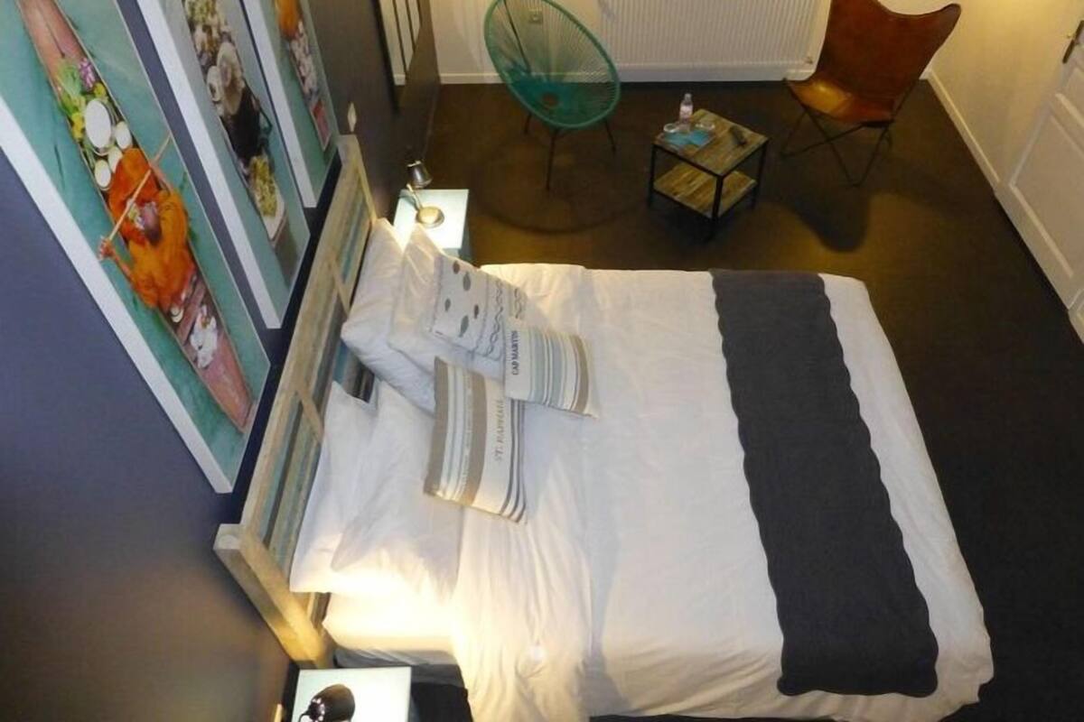 A neatly made bed with a striped blanket and pillows, a small round table with a bottle and glasses, a green chair, and a radiator in a room with a window.