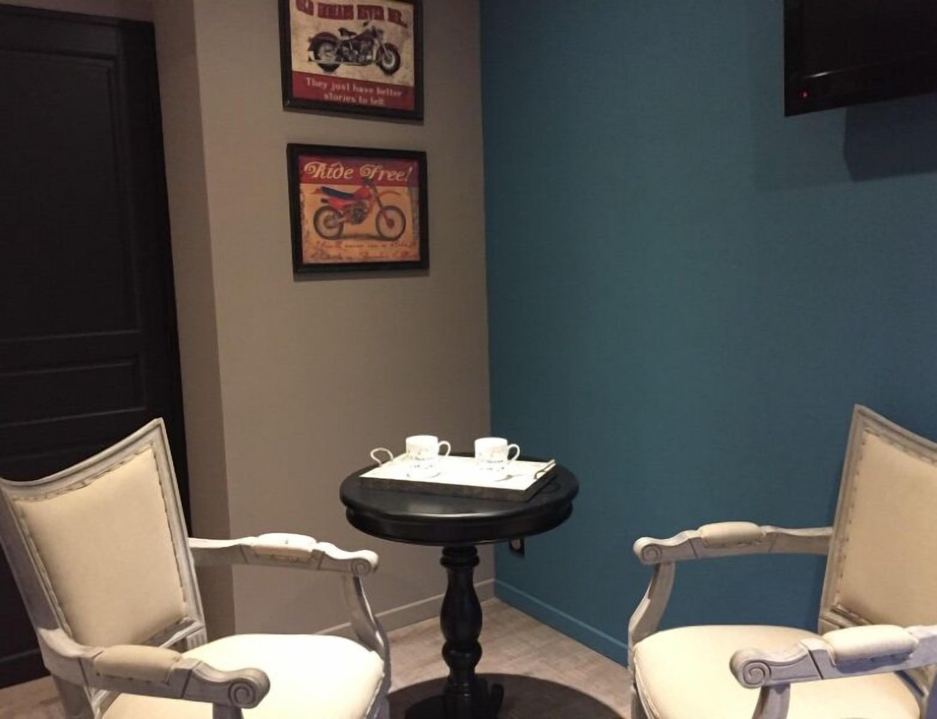 A room with two white upholstered chairs, a small round table with a black base, and two white cups. There are two framed posters on the wall, one with a motorcycle and the other with the text "Ride Free!" The room has a blue accent wall and a black door.
