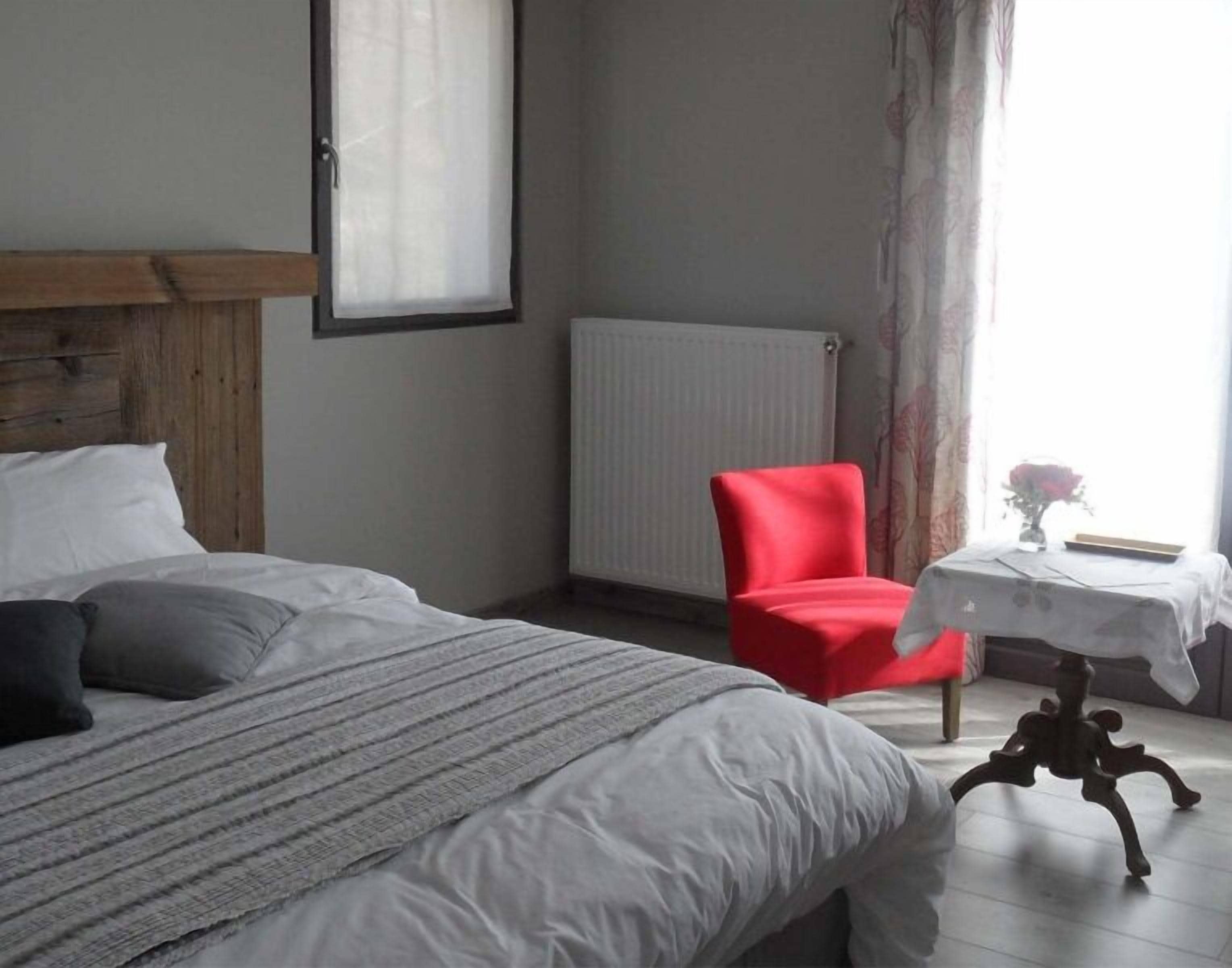 Une chambre à coucher avec un lit, une chaise rouge, une petite table avec un vase et une fenêtre avec des rideaux.