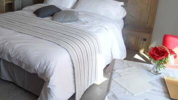 Un lit bien fait, avec du linge de lit blanc et une couverture grise, une tête de lit en bois et une petite table sur laquelle est posé un vase rempli de fleurs rouges.