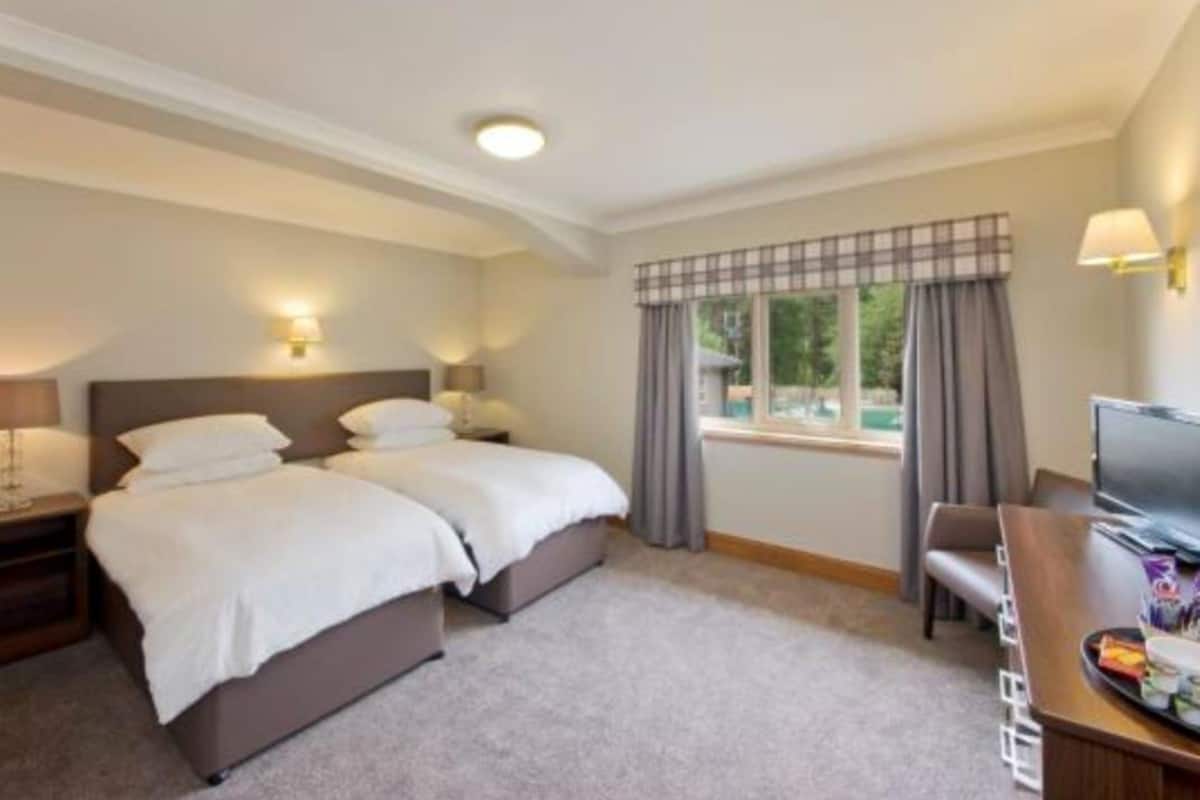 A hotel room with two beds, a desk with a TV, and a window with curtains.
