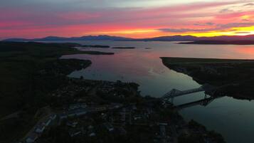 An aerial view of a coastal town with a bridge, calm water, and distant mountains during sunset.