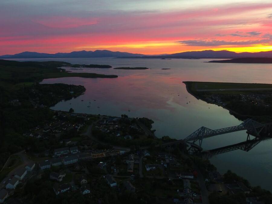 An aerial view of a coastal town with a bridge, calm water, and distant mountains during sunset.