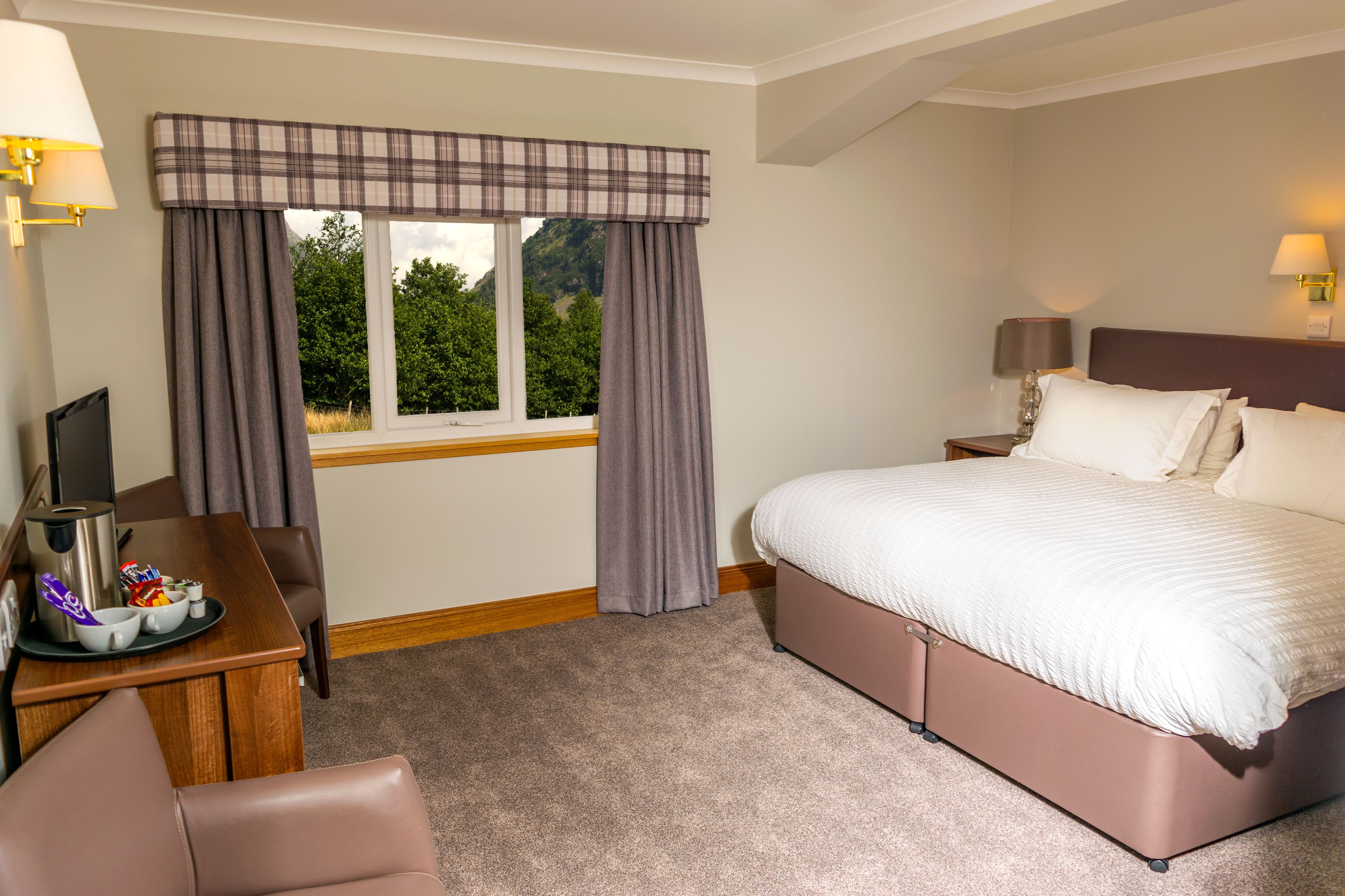 A hotel room with a bed, a desk with a coffee maker, a chair, and a window with curtains.