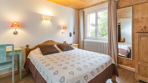 Une chambre avec un plafond en bois, un lit recouvert d’une couvre-lit à motifs, deux lampes de chevet, une fenêtre avec des rideaux et une armoire en bois.