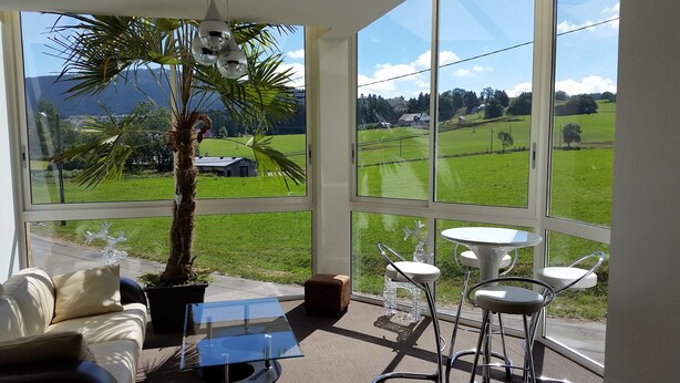 Une véranda avec une table en verre, un canapé et un fauteuil. Elle est dotée d’une grande fenêtre offrant une vue sur un champ verdoyant et une montagne.
