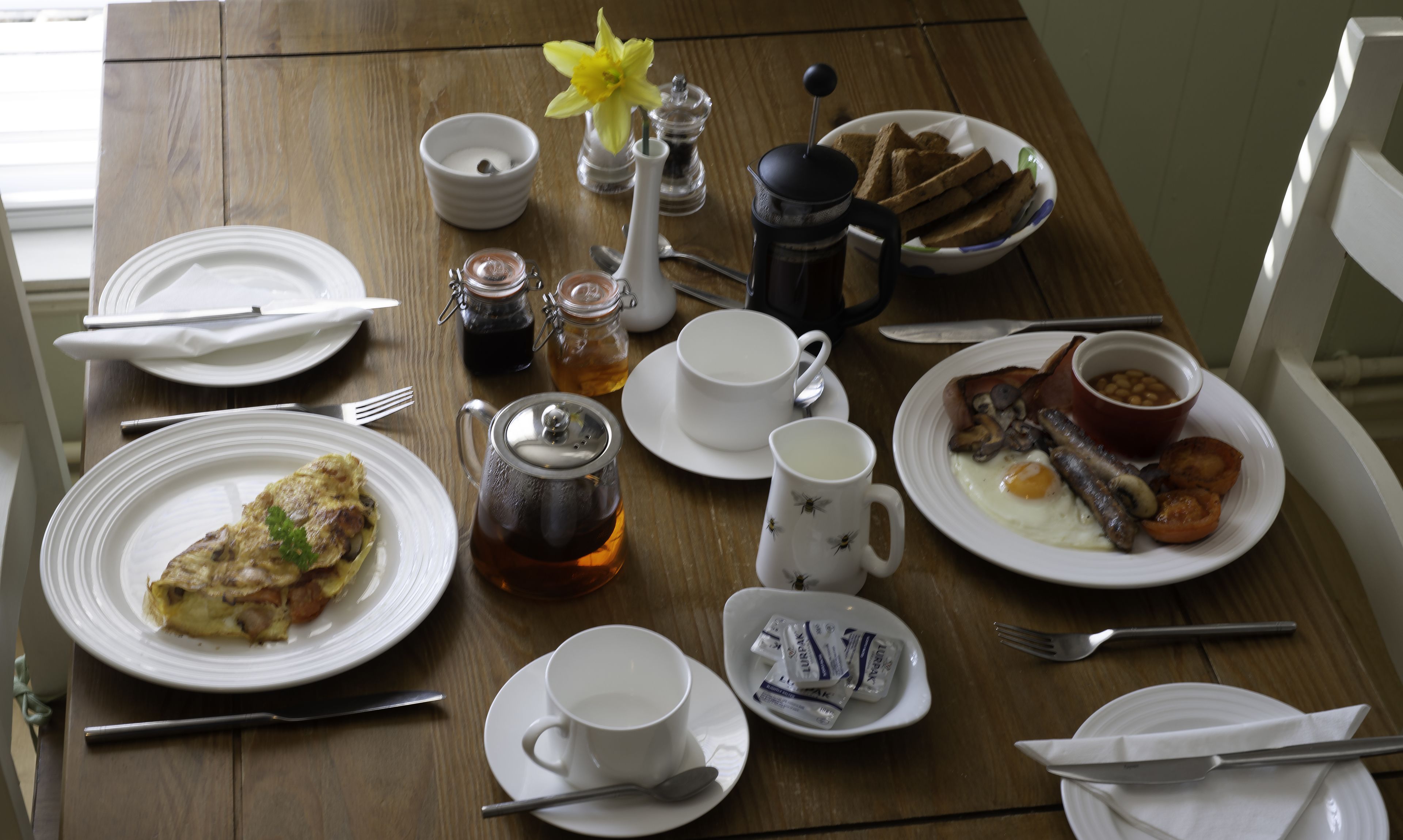 Une table de petit-déjeuner avec des assiettes remplies de nourriture, des tasses, une théière et un bouquet de fleurs.