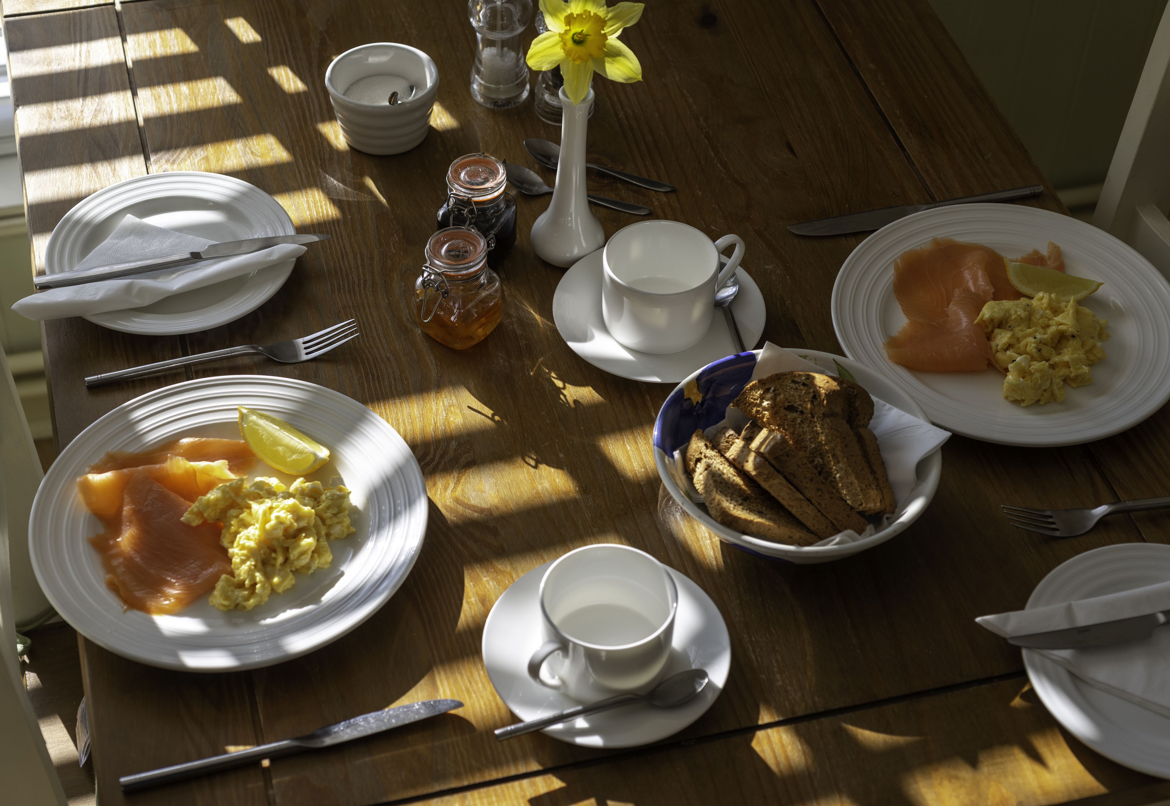A wooden table with breakfast items including eggs, smoked salmon, toast, and a flower vase.