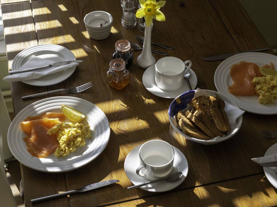 A wooden table with breakfast items including eggs, smoked salmon, toast, and a flower vase.