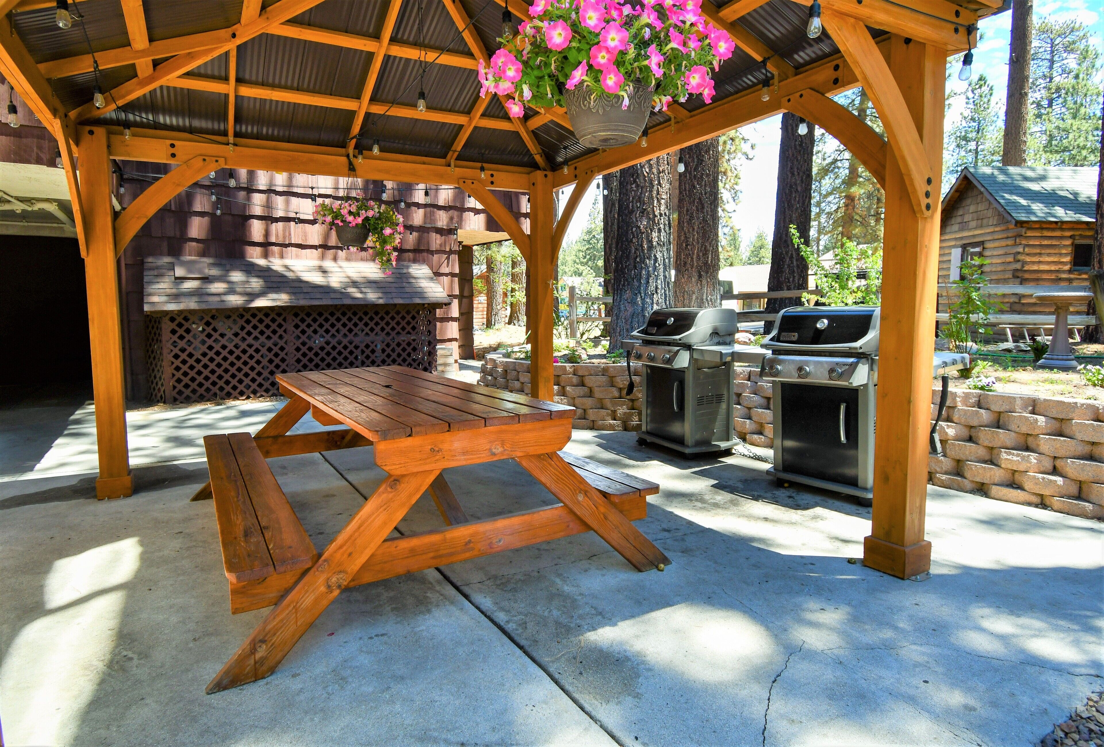 A wooden gazebo with a picnic table, barbecue grills, and hanging flower baskets.