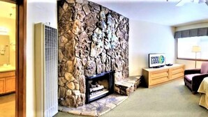 Habitación de hotel con chimenea de pared de piedra, televisor, cama, sillón y ventana con vistas.